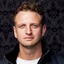 Portrait of a young man with short curly hair and blue eyes wearing a black jacket over a white shirt against a dark patterned background.