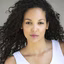 Portrait of a woman with long curly hair wearing a white sleeveless top against a neutral background.